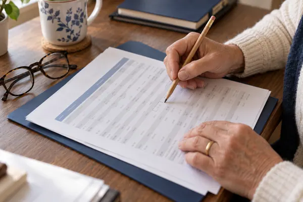 Hands using a pencil to mark a row on a printed report beside reading glasses.